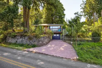 A long, gently sloped paved driveway leads to the understory garage/hobby are to total 3 covered parking spaces. The terraced gardens step up to the courtyard on the right as well as stairs on the left leading to the shop.