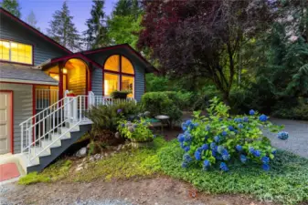 Twilight view of the front entry framed by lush landscaping and vibrant hydrangeas—showcasing warm curb appeal and inviting design.