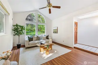 Bright living room with soaring vaulted ceilings, a statement arched window, and ceiling fan—framing peaceful views of the surrounding evergreens.