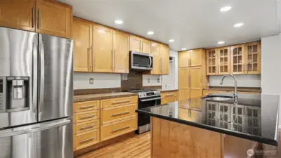 Modern kitchen with natural toned cabinetry. Spacious island features a sink, dishwasher, outlet, and plenty of storage—perfect for meal prep and casual gatherings.
