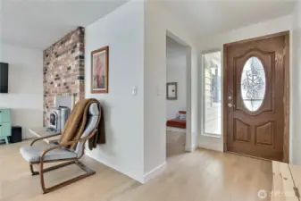 Step inside to a light and welcoming entry with beautiful new wide-plank hardwood laminate flooring and a decorative glass-inset front door that adds charm and character. Natural light pours in from the adjacent sidelight window, highlighting the fresh interior paint and clean, modern lines. A cozy peek into the front sitting room and a glimpse of the brick-accented fireplace create a feeling of home right from the start.