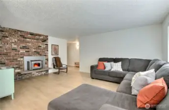 Spacious and light-filled living room features stunning wide-plank laminate flooring and a cozy wood-burning fireplace with a striking full brick surround. The open layout offers plenty of room for entertaining or relaxing, with large windows bringing in natural light. A warm and welcoming centerpiece to the home!