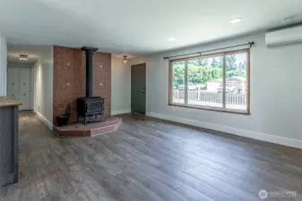 Family room looking back to front door showing wood stove and mini-split system. Natural lighting with big windows.