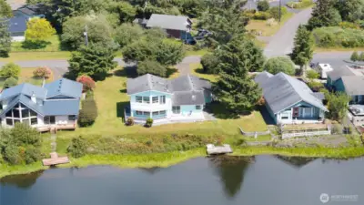 This bird's-eye view captures the property's exceptional canal-front setting — a charming two-story home with a wraparound deck, lush green yard, and private dock right on the water's edge, perfectly positioned to enjoy the tranquil canal and the best of coastal Washington living.