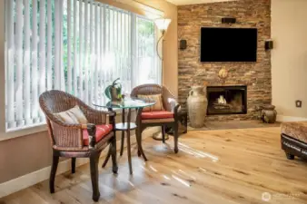 Highlighted by a stunning floor-to-ceiling stacked stone fireplace and mounted TV, perfect for cozy evenings at home. Natural light pours through the oversized window with vertical blinds, creating a bright and airy atmosphere. The elegant bistro nook offers a charming spot for morning coffee or intimate conversation, framed by lush outdoor greenery.