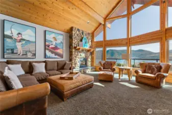 View of of living room w/ oversized windows, river rock fireplace and pine ceilings.