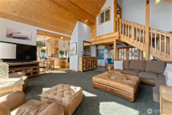 View 2 of of living room w/ oversized windows, river rock fireplace and pine ceilings, looking towards the entryway, kitchen and dining room and stairs to the upper level bedroom/bunk room w/ bath.