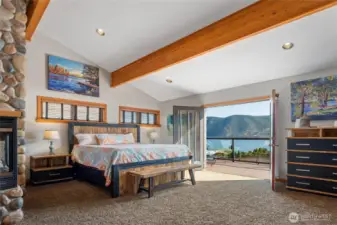 View of main level Primary Bedroom suite w/ stone fireplace and French Doors leading to the lake view deck seating area w/ glass railings.