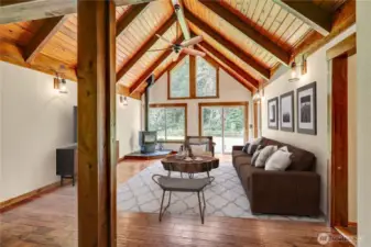 Spacious living area with high, vaulted ceilings made of warm-toned wood planks and exposed wooden beams. A ceiling fan hangs from the peak. The room features a wood stove and large windows and glass doors that look out onto a natural, wooded setting. Virtually staged