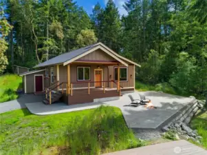 Charming island retreat surrounded by evergreens, featuring a welcoming front porch, spacious patio with firepit, and modern finishes for easy indoor-outdoor living.