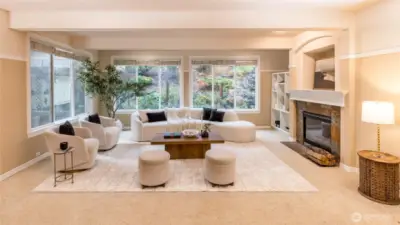 A spacious and serene living room featuring large picture windows overlooking the landscaped backyard. The room includes a stone-accented fireplace, built-in shelving, and generous seating areas—ideal for relaxing or entertaining in style.