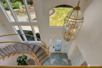 From the second floor, look down into a striking entryway that showcases the elegant spiral staircase and soaring ceilings. Large windows frame serene views of the front grounds, filling the space with natural light and a sense of openness.