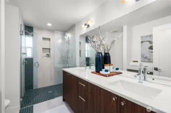 Photos of model home 9213 with similar finishes & floor plan. The primary bath gives a spa like plcae: dual sinks, a walk-in shower, warm tones, and heated floors for extra comfort.