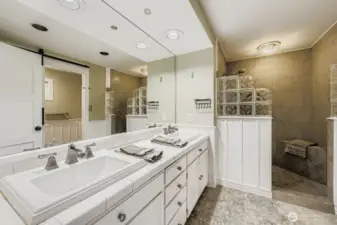 The well-designed Primary bath boasts a large walk-in tiled shower with glass blocks, dual sinks, excellent lighting, abundant cabinetry, tile countertops and flooring.