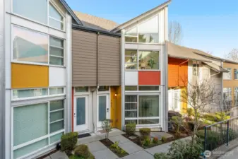 This vibrant Element townhome's exterior features modern multi-toned siding in white, gray, orange, and yellow, large glass windows, private entry doors with sidelights, and a tidy front patio with low-maintenance landscaping.