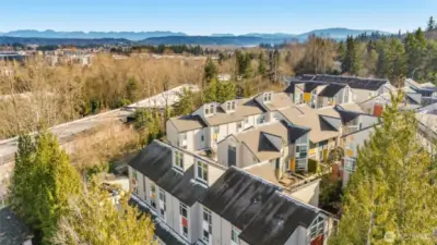 This panoramic aerial view captures a vibrant townhome community nestled amid lush evergreens, with colorful modern units featuring peaked roofs, surrounded by dense forest and distant snow-capped mountain horizons under a clear blue sky.
