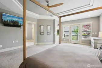 Expansive primary bedroom suite featuring plush carpeting, tray ceiling, ceiling fan, and abundant natural light. French doors provide direct access to the private deck, creating a seamless connection to outdoor living and enhancing the overall sense of space and comfort.