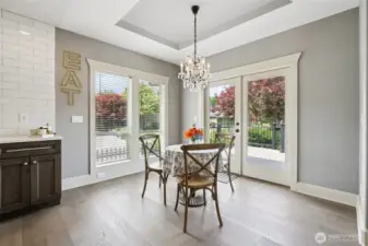 Light-filled dining area featuring tray ceiling detail, statement chandelier, and direct access to the outdoor deck through French doors. Large windows provide natural light and views of the landscaped yard, creating a seamless flow between indoor dining and outdoor living.