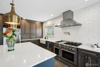 Well-appointed gourmet kitchen featuring custom appliance cabinetry, expansive quartz countertops, and a professional-grade double oven range with six burners and center griddle. Additional highlights include a statement vent hood, farmhouse sink, generous island seating, and thoughtful storage throughout, creating a highly functional space designed for both everyday use and entertaining.