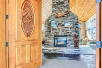 Unique custom carved wood entry doors lead to a slate entry & impressive stone hearth on the double-sided propane fireplace