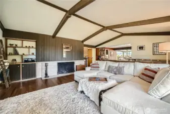 Mid-century warmth meets modern comfort in this inviting living room featuring vaulted ceilings with exposed beams, rich wood-paneled accents, and a classic wood-burning fireplace. Built-in shelving adds character and functionality, while the open flow to the dining and kitchen areas creates the perfect setting for entertaining or relaxing in style. A cozy, elevated vibe that feels like home the moment you walk in.