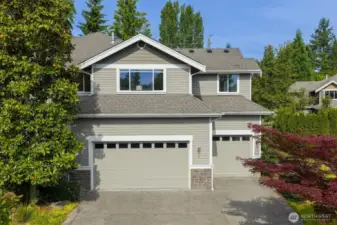 This side of the home showcases a spacious 3-car garage with two distinct doors—one double and one single—framed by crisp white trim and topped with transom-style windows that add architectural interest and natural light. The garage exterior features a combination of horizontal siding and stone accents at the base. The driveway is wide and cleanly finished, offering ample space for parking and easy access.