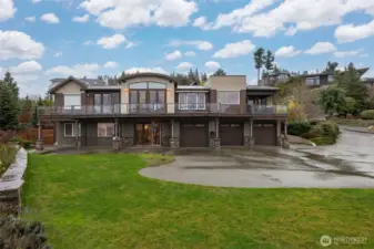 The rear elevation of the home showing the wraparound deck and architecturally  designed features of the home. There is an abundance of parking area that is convenient for turning full-sized trucks and SUVs around, too.