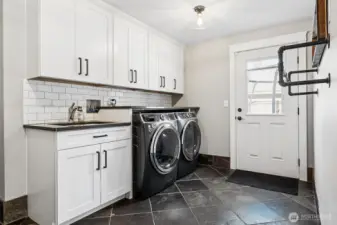 Located just off the garage, the laundry room doubles as a true utility hub with a newer washer and dryer that stay, plus a generous folding counter right above to keep things efficient. The utility sink gets a nice upgrade with a full-height subway tile backsplash that runs cleanly up to the upper cabinets, giving it a polished, finished feel. White shaker cabinetry provides plenty of storage to keep supplies tucked away, while durable tile flooring handles wet shoes, laundry days, and real life without complaint. Across the room, the industrial-style pipe hanging system adds a cool, functional touch for air drying or staging clothes. There’s also a door out to the side yard with a window that brings in light and adds easy access, making this space feel more like a smart mudroom than just a laundry stop. There is also a fridge and deep freezer in the garage that can stay with the home.