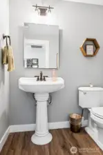 This powder room features a pedestal sink and beveled mirror that give it a traditional touch, while the bronze fixture adds a bit of warmth and contrast. The same wide plank luxury vinyl flooring runs through here, keeping the flow uninterrupted and cohesive with the rest of the home. Soft dove gray walls paired with white trim continue the interior palette, creating a space that feels light, balanced, and thoughtfully consistent from room to room.