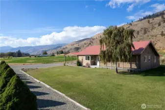 Beautiful side view of the home with covered porch, established trees, and wide-open green space leading to mountain views.