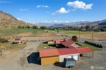 Side aerial perspective highlighting the home, carport, barn-style outbuildings, and ample gravel parking.