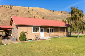 Welcoming front view of the home with covered porch, mature tree, and a metal roof set against a mountain backdrop.