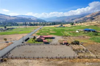 Sweeping aerial view of the property featuring the large fenced riding arena, outbuildings, and lush surrounding fields with mountain vistas.