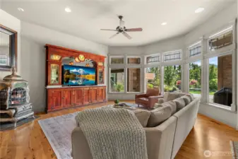 Light-filled living room featuring expansive windows with transom accents that frame beautiful outdoor views. Warm hardwood floors, high ceilings, and a cozy stove create an inviting space for relaxing or entertaining.