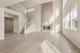 Stunning wide-angle view of the great room featuring soaring two-story ceilings, an open staircase with white painted wood railing, a modern linear electric fireplace, and oversized windows bathing the space in beautiful natural light. Photos are representative only and may display different color package and/or upgrades.