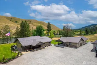 A stunning riverside property featuring two beautifully designed single-level homes with modern metal roofs and stone accents. Nestled against rolling hills and lush greenery, the residences are connected by concrete sidewalks and surrounded by an expansive gravel courtyard. The serene landscape, complete with mature trees and a nearby river, creates the perfect blend of comfort, privacy, and natural beauty.