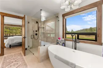 This beautifully designed primary bathroom features a modern-rustic aesthetic with natural wood accents and luxurious amenities. The space includes a frameless glass walk-in shower with matte black fixtures, a freestanding soaking tub positioned beside a large picture window that offers stunning views of a serene meadow and mountains. Natural light pours in, highlighting the light neutral tile flooring and walls.
