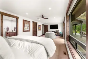 A view from inside the primary bedroom highlights its warm and inviting design, featuring rich wood trim, doors, and baseboards that add a rustic elegance. The king-sized bed is complemented by simple yet tasteful furnishings, including a vintage-style lamp and a wooden trunk at the foot of the bed. Artwork along the wall and recessed lighting create a cozy, lived-in ambiance, while the open floor plan enhances the spacious feel.