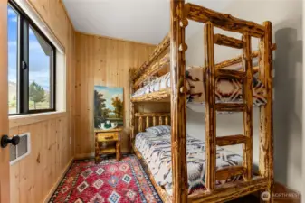 A cozy bunk room with warm wood paneling, rustic log-frame bunks, and a colorful accent rug — perfect for hosting family or guests in comfort and style.