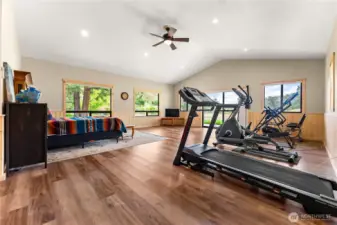 An expansive bonus room doubles as a home gym and guest suite, offering space for fitness equipment, a bed, and a seating area. Large windows fill the room with natural light while showcasing serene river views.