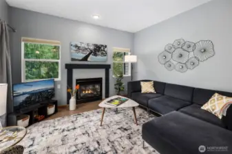 Bright and welcoming living room centered around a cozy gas fireplace—perfect for relaxing or entertaining. Large windows and sliding doors open to a private deck surrounded by lush green trees, blending indoor comfort with outdoor serenity