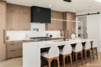 A double-waterfall quartz island anchors the kitchen, ideal for gathering and entertaining.  Custom oak cabinetry and a commanding matte black range hood add elegance and drama.