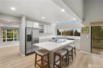 Open, sunlit kitchen with expansive island, seating, skylight, white cabinetry, stainless appliances, wide plank waterproof floors, and  an inviting entertaining layout.