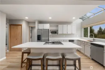 Stylish open kitchen with oversized island, seating, skylight, white cabinetry, stainless appliances, wide plank floors, and seamless entertaining flow design.