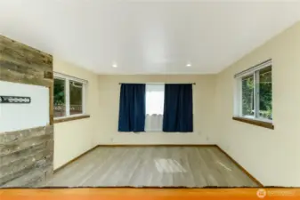 Spacious living area filled with natural light from multiple windows, offering a bright and welcoming atmosphere. The room features updated wood-look flooring, recessed lighting, and a rustic accent wall with built-in TV mount for added character. Neutral walls provide a versatile backdrop, ready for personal touches.