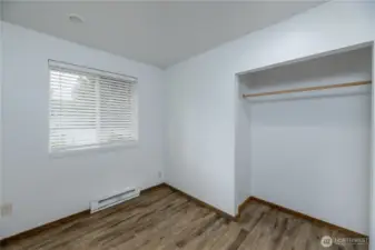 Cozy bedroom featuring durable wood-look flooring, a large window with blinds providing natural light, and a spacious open closet ready for customization.