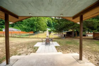 Covered patio overlooking a spacious, fully fenced yard with mature trees for added privacy. A paved patio area provides the perfect spot for outdoor dining and entertaining, complete with space for a table and chairs. The large open yard offers plenty of potential for gardening, play, or pets, making it a versatile extension of the home’s living space.