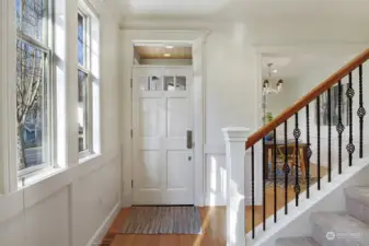 Sunlit entryway with warm oak floors, wrought iron spindle staircase and a wall of windows.