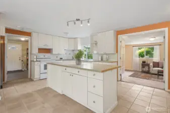 The kitchen keeps it classic with white shaker-style cabinets, silver hardware, and a clean tile backsplash. White appliances blend in, while a corner sink sits under a garden shelf window that’s basically begging for fresh herbs. The back door carries the same arched glass detail as the front, and through the French doors you’ve got flex space that works as a third bedroom or whatever you need.