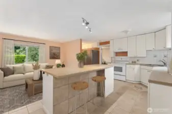 This view shows how the kitchen stays bright and airy, with light bouncing off white finishes and oversized windows. The bar counter is finished with protective tile underneath, so stool scuffs never stand a chance. Across from the island, another cadet wall heater lets you dial in the temperature room by room, and blinds are already in place when you want to control the light.